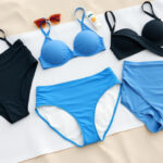 Naisten bikinialaosia shortsityylillä ja lantiolla istuvia malleja aseteltuna ranta-atmosfääriin hiekalle aurinkovoidetta ja aurinkolasia viereen.